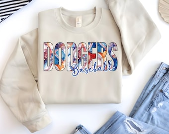 May include: A cream-colored sweatshirt with the word "Dodgers" in large, colorful letters, with baseballs and watercolor designs. Below the word "Dodgers" is the word "Baseball" in a blue script font. The sweatshirt is folded.
