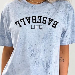 May include: A light blue t-shirt with a white tie-dye pattern. The shirt has black text that reads "BASEBALL LIFE" in a varsity-style font.