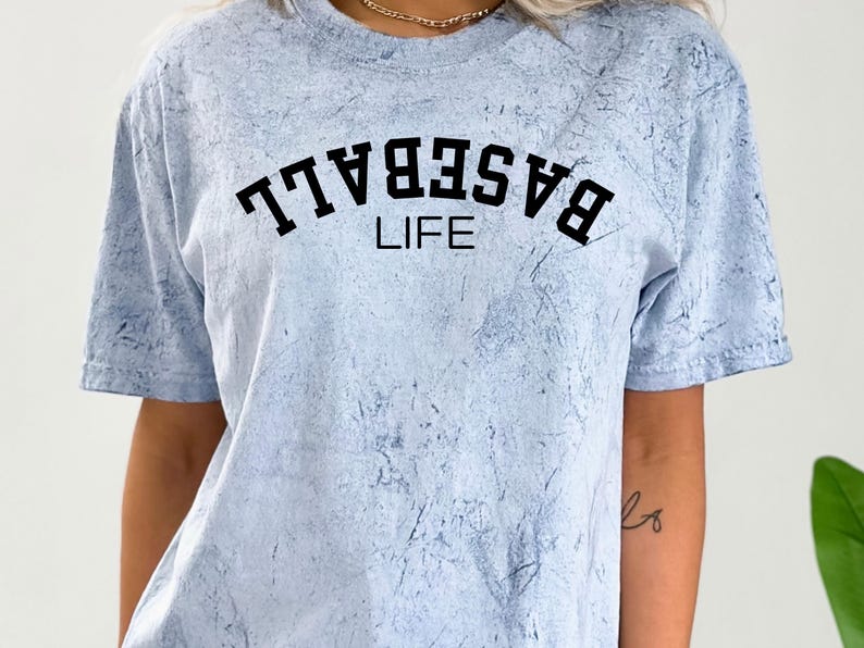 May include: A light blue t-shirt with a white tie-dye pattern. The shirt has black text that reads "BASEBALL LIFE" in a varsity-style font.
