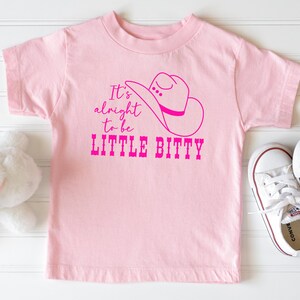 May include: Pink t-shirt with a pink cowboy hat graphic and the text "It's alright to be LITTLE BITTY".