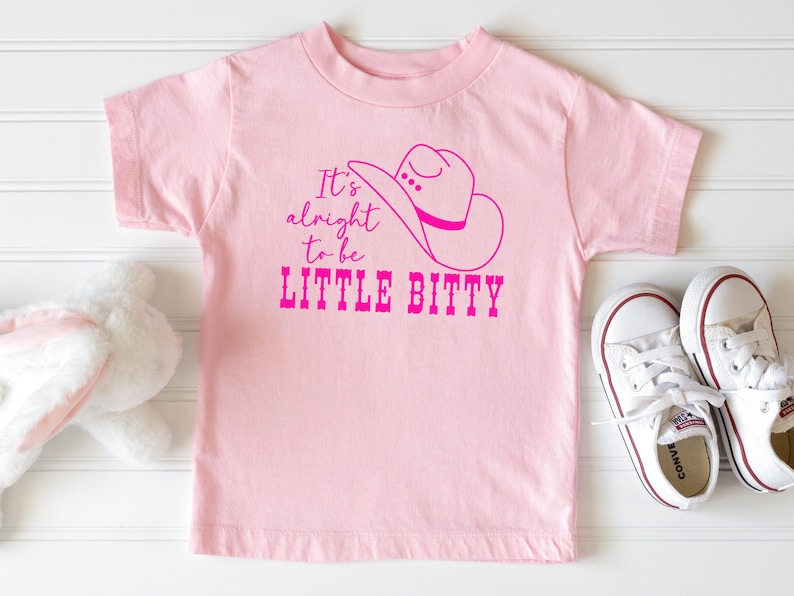 May include: Pink t-shirt with a pink cowboy hat graphic and the text "It's alright to be LITTLE BITTY".