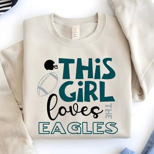 Può includere: Una felpa color crema con la scritta "THIS GIRL loves THE EAGLES" in verde acqua, nero e grigio. Un casco da football nero e un pallone da football grigio sono presenti sulla maglietta. La felpa è piegata con cura.