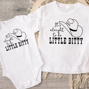 May include: Two white short-sleeved shirts with black text and a cowboy hat graphic. The text on the shirts reads "It's alright to be little bitty".
