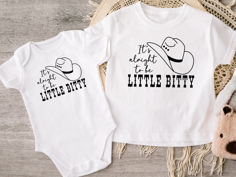 May include: Two white short-sleeved shirts with black text and a cowboy hat graphic. The text on the shirts reads "It's alright to be little bitty".