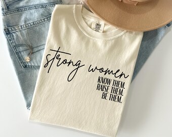 May include: Cream-colored t-shirt with the words "strong women" in a script font. Below, the text continues with "KNOW THEM. RAISE THEM. BE THEM." The shirt is laid on top of blue jeans, with a necklace and a tan hat.
