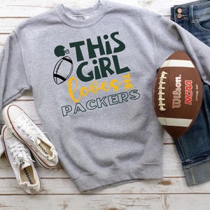 May include: A gray sweatshirt with the text "THIS GIRL LOVES PACKERS" in green and yellow, along with a football graphic. A brown football, white sneakers, and blue jeans are also in the image.