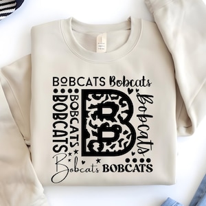 May include: A beige sweatshirt with the word "BOBCATS" in black lettering. The letter "B" is in the center, filled with a leopard print design. The word "Bobcats" is repeated around the letter "B".