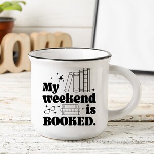 May include: White ceramic mug with a black rim and handle. The mug has a black graphic that says "My weekend is BOOKED." with a stack of books and glasses.