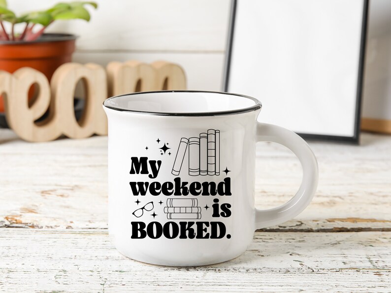 May include: White ceramic mug with a black rim and handle. The mug has a black graphic that says "My weekend is BOOKED." with a stack of books and glasses.