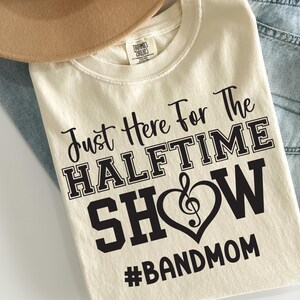 May include: Cream-colored t-shirt with the text "Just Here For The HALFTIME SHOW #BANDMOM" in black. The word "SHOW" is inside a heart with a treble clef. The shirt is folded on top of light blue jeans and a tan hat.