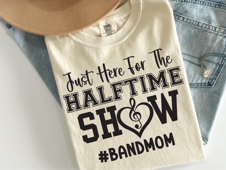 May include: Cream-colored t-shirt with the text "Just Here For The HALFTIME SHOW #BANDMOM" in black. The word "SHOW" is inside a heart with a treble clef. The shirt is folded on top of light blue jeans and a tan hat.