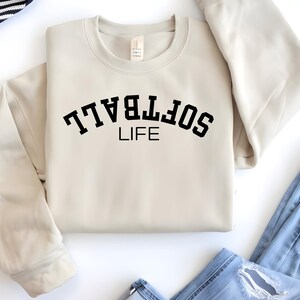 May include: A cream-colored sweatshirt with the words "SOFTBALL LIFE" printed in black, arched across the chest. The sweatshirt is folded, with the sleeves neatly arranged. Blue jeans are visible in the bottom right corner.