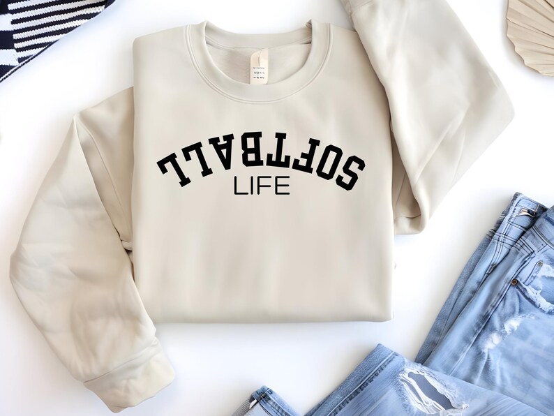 May include: A cream-colored sweatshirt with the words "SOFTBALL LIFE" printed in black, arched across the chest. The sweatshirt is folded, with the sleeves neatly arranged. Blue jeans are visible in the bottom right corner.