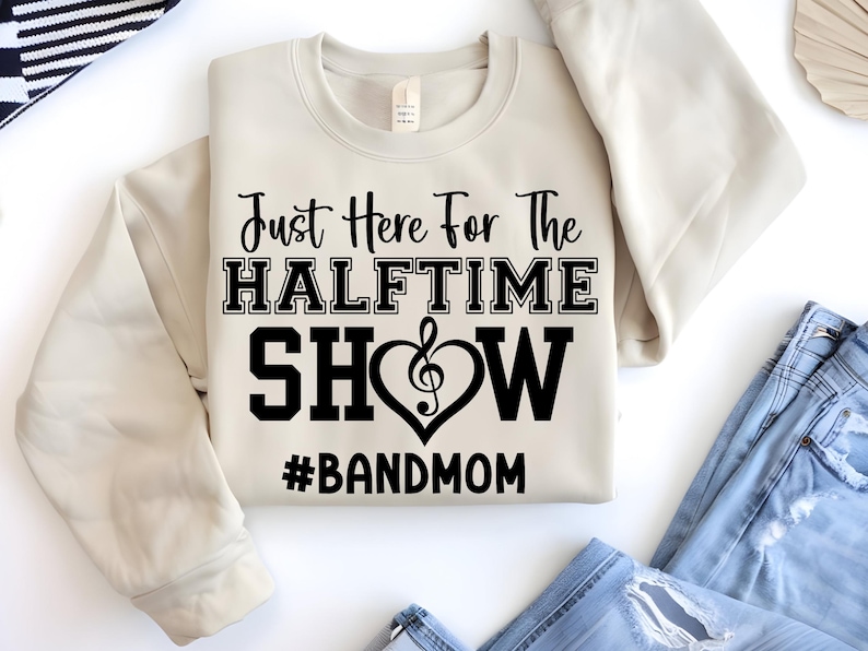 May include: A cream-colored sweatshirt with the text "Just Here For The HALFTIME SHOW" in black block letters. A treble clef is inside a heart shape. The hashtag "#BANDMOM" is below the text.