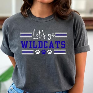 May include: Dark gray t-shirt with the text "Let's go WILDCATS" in white and blue, along with paw print graphics. The shirt has a crew neck and short sleeves, suitable for casual wear.
