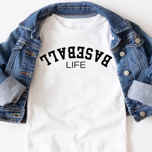 May include: White t-shirt with black text that reads "BASEBALL LIFE" on a white t-shirt. The t-shirt is folded and displayed on a denim jacket.