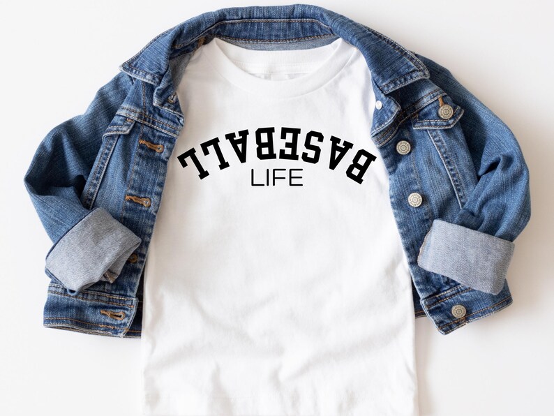 May include: White t-shirt with black text that reads "BASEBALL LIFE" on a white t-shirt. The t-shirt is folded and displayed on a denim jacket.