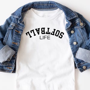 May include: White t-shirt with the text "SOFTBALL LIFE" in black, displayed under a blue denim jacket. The shirt's design is centered, and the jacket is neatly draped around it, creating a casual, sporty look.