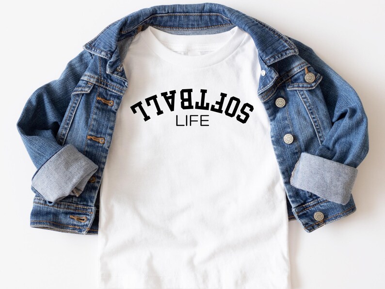 May include: White t-shirt with the text "SOFTBALL LIFE" in black, displayed under a blue denim jacket. The shirt's design is centered, and the jacket is neatly draped around it, creating a casual, sporty look.