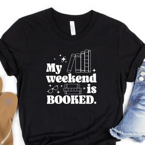 May include: Black t-shirt with white text that reads "My weekend is BOOKED."  The text is in a retro font and is accompanied by a graphic of a stack of books and a pair of glasses.