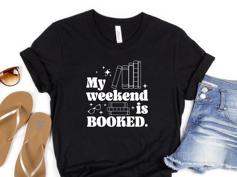 May include: Black t-shirt with white text that reads "My weekend is BOOKED."  The text is in a retro font and is accompanied by a graphic of a stack of books and a pair of glasses.
