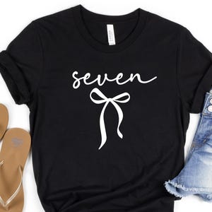 May include: Black t-shirt with the word "seven" in a cursive font and a white bow graphic. The shirt is paired with denim shorts and sandals, suggesting a casual, everyday style.