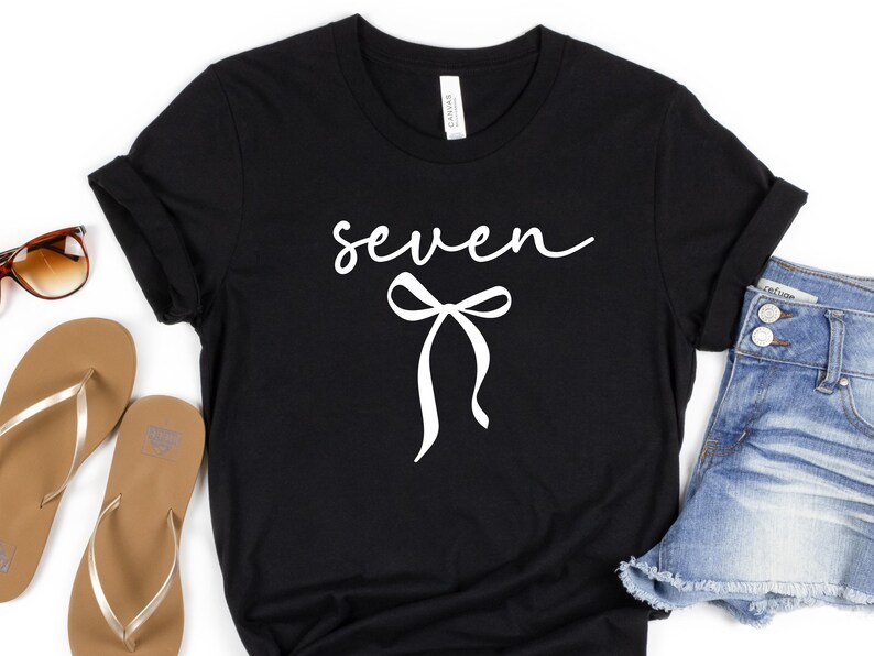 May include: Black t-shirt with the word "seven" in a cursive font and a white bow graphic. The shirt is paired with denim shorts and sandals, suggesting a casual, everyday style.