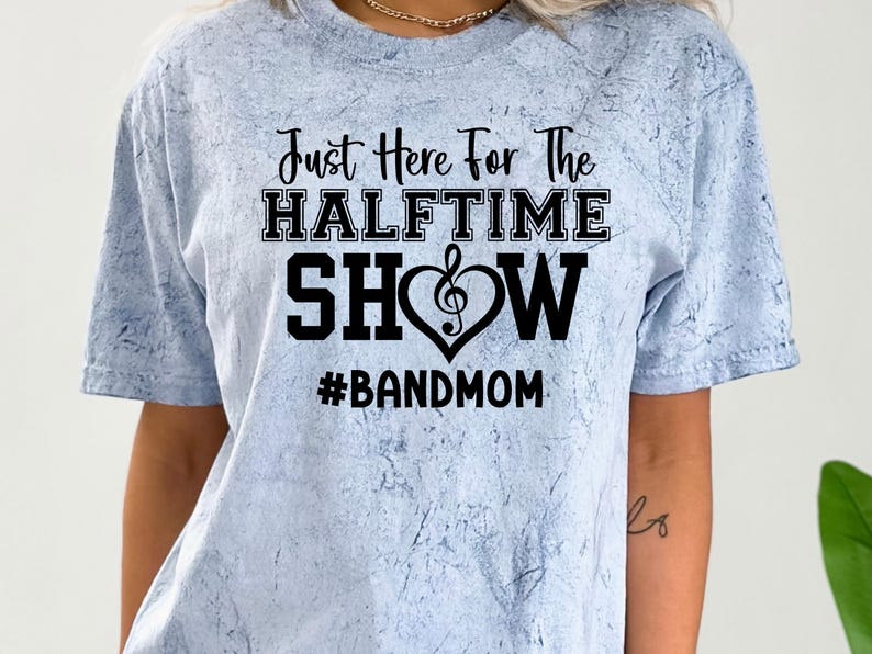 May include: Light blue tie-dye t-shirt with the text "Just Here For The Halftime Show" in black. A musical note heart design is between "Show" and "Halftime". The hashtag "#BANDMOM" is below the text.