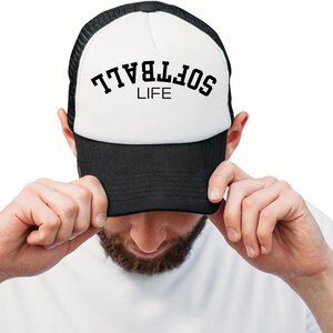 May include: A black and white trucker hat with the word "SOFTBALL" arched across the top in black letters, with "LIFE" underneath. The hat is being held by a person.