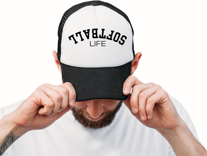 May include: A black and white trucker hat with the word "SOFTBALL" arched across the top in black letters, with "LIFE" underneath. The hat is being held by a person.