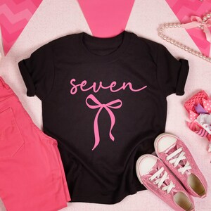 May include: Black t-shirt with the word "seven" in pink script and a pink bow graphic. The image also includes pink pants, pink glitter sneakers, a pink flower, and a decorative bow.