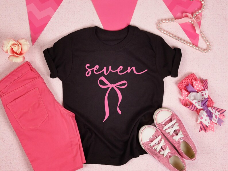May include: Black t-shirt with the word "seven" in pink script and a pink bow graphic. The image also includes pink pants, pink glitter sneakers, a pink flower, and a decorative bow.