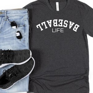 May include: A dark grey t-shirt with white text that reads "BASEBALL LIFE" in an arc. The shirt is paired with blue jeans and black trainers.