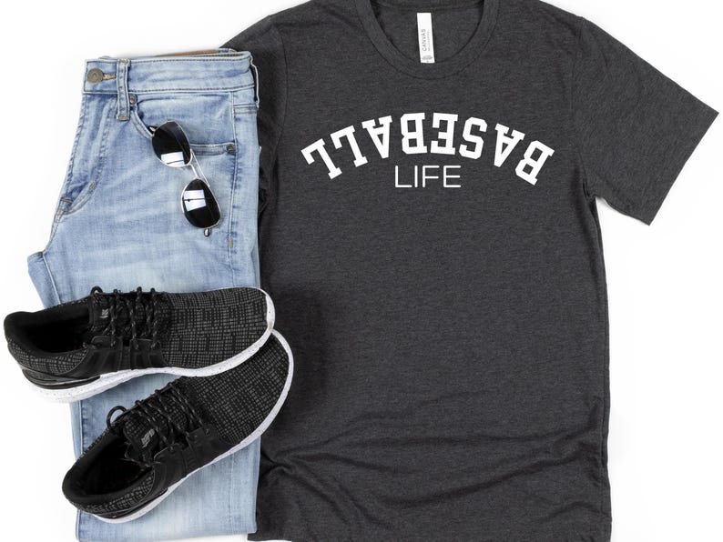 May include: A dark grey t-shirt with white text that reads "BASEBALL LIFE" in an arc. The shirt is paired with blue jeans and black trainers.