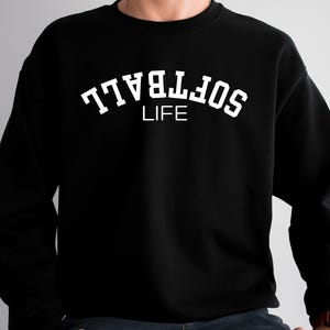 May include: Black sweatshirt with the white text "SOFTBALL LIFE" in an arched design. The sweatshirt is a crew neck style and appears to be made of a soft material. The text is in a bold, sans-serif font.