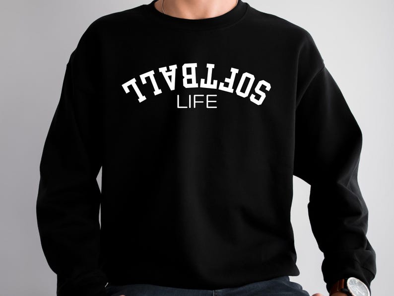 May include: Black sweatshirt with the white text "SOFTBALL LIFE" in an arched design. The sweatshirt is a crew neck style and appears to be made of a soft material. The text is in a bold, sans-serif font.