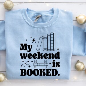 May include: Light blue sweatshirt with the text "My weekend is BOOKED." in black. The text is in a retro font and is accompanied by a stack of books and glasses.