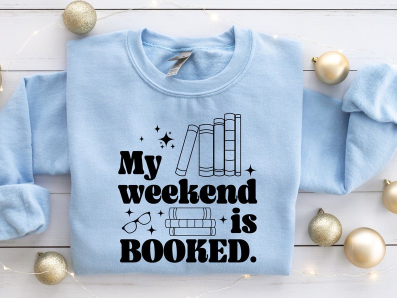 May include: Light blue sweatshirt with the text "My weekend is BOOKED." in black. The text is in a retro font and is accompanied by a stack of books and glasses.