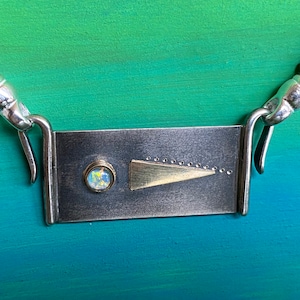May include: A silver necklace with a rectangular pendant featuring a gold triangle and a small opal gemstone. The necklace is made of silver beads and has a silver clasp.