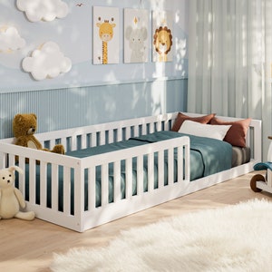 Op de afbeelding: Wit kinderbed met een wit veiligheidshek aan één kant. Het bed is opgemaakt met een blauwe en witte beddengoedset. Er liggen knuffels op de vloer naast het bed.