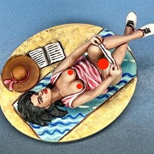 May include: A painted figurine of a woman lying on a beach towel, wearing a striped shirt and white shoes, reading a book. She is on a beach scene with a straw hat and a book.