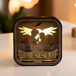 May include: A wooden Final Fantasy VIII night light. The square-shaped light features a silhouette of two figures with angel wings, illuminated by a warm glow. The text "Final Fantasy VIII" is displayed in a gold color.