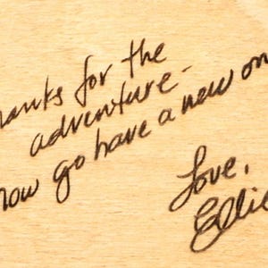 May include: A handwritten message in black ink on a light brown wooden surface. The text reads "Thanks for the adventure - now go have a new one! Love, Ellie."