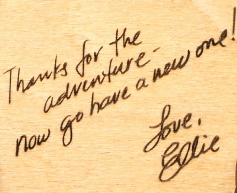 May include: A handwritten message in black ink on a light brown wooden surface. The text reads "Thanks for the adventure - now go have a new one! Love, Ellie."