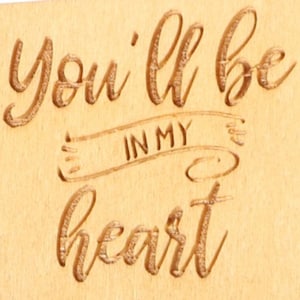 May include: A wooden plaque with the engraved phrase "You'll be in my heart." The text is in a flowing script and is centered on the light-colored wood. The words "IN MY" are within a banner design.
