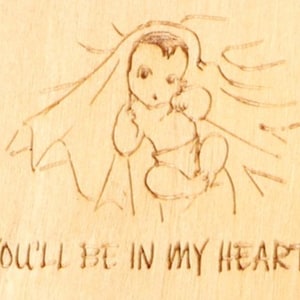 May include: A light-colored wooden plaque with a sketched image of a baby and the text "YOU'LL BE IN MY HEART" etched below. The baby is depicted with a halo and wrapped in a cloth.