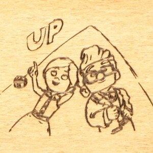May include: A wood-burned image featuring characters from the animated film "Up." The image depicts two cartoon figures under a drawn roof, with the word "UP" above them. The characters are drawn in a simple, illustrative style.