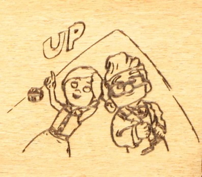 May include: A wood-burned image featuring characters from the animated film "Up." The image depicts two cartoon figures under a drawn roof, with the word "UP" above them. The characters are drawn in a simple, illustrative style.