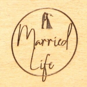 May include: A wooden surface with a circular design etched into it. The design features the words "Married Life" in a cursive font, enclosed within a circle. Above the text, there is a silhouette of a couple embracing.