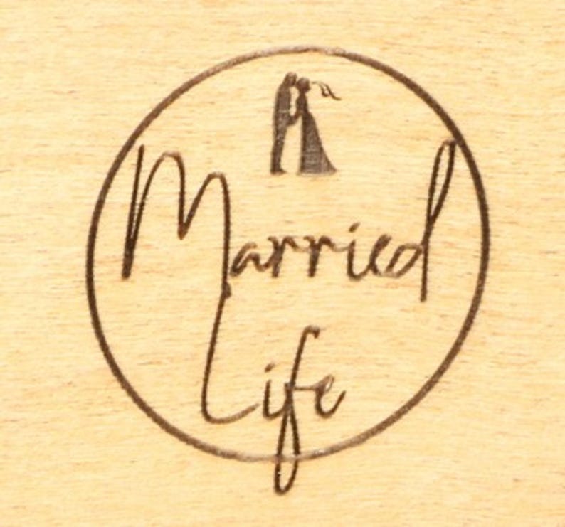 May include: A wooden surface with a circular design etched into it. The design features the words "Married Life" in a cursive font, enclosed within a circle. Above the text, there is a silhouette of a couple embracing.